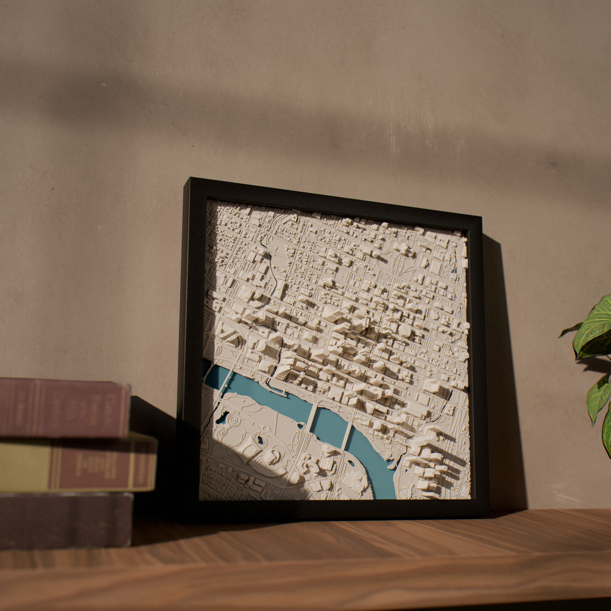A framed 3D city map wall art piece with textured off-white buildings and a winding turquoise river, displayed on a wooden shelf next to stacked books against a warm beige wall.