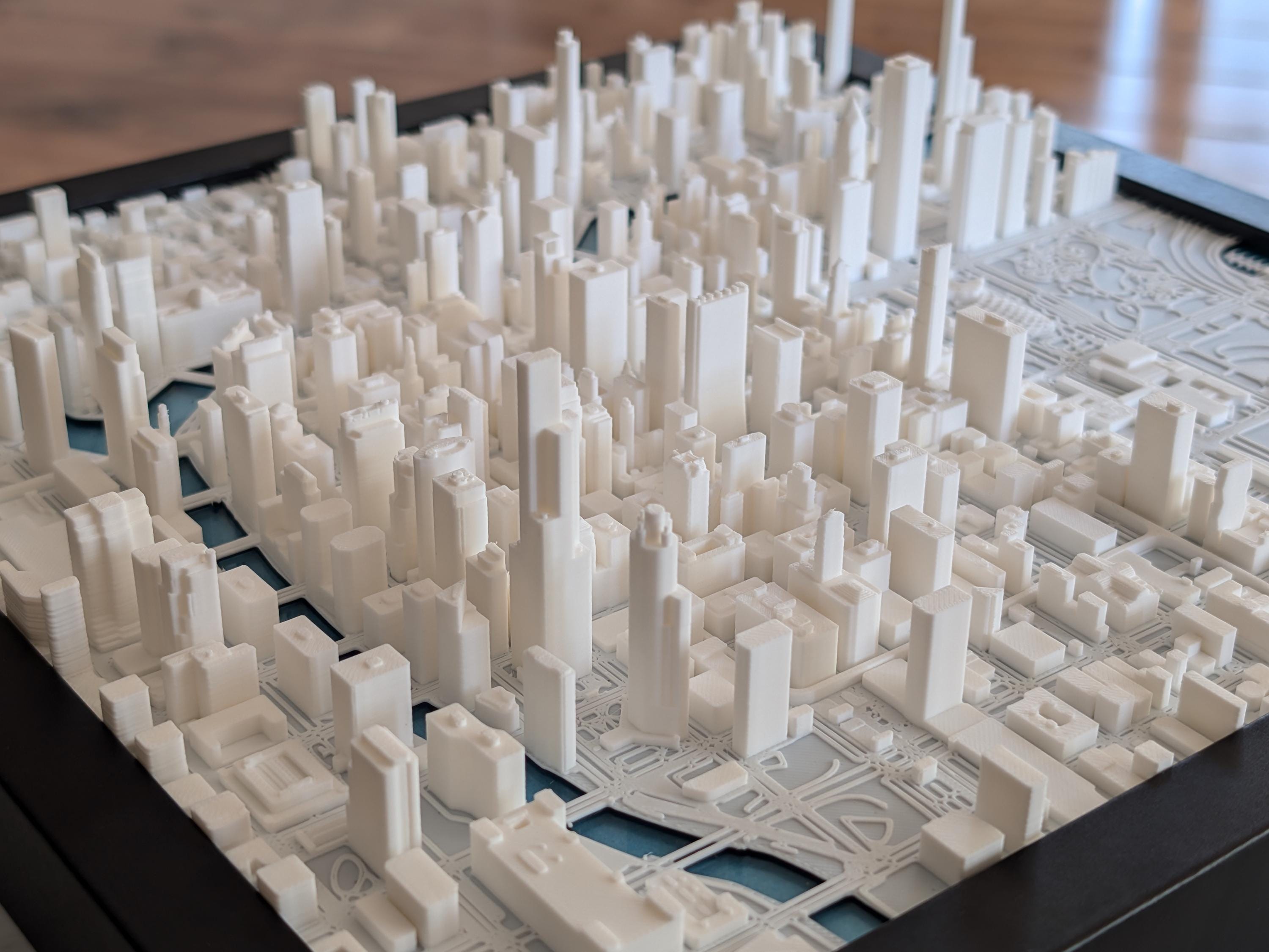 The Chicago 3D Map is a detailed white cityscape model with buildings of different heights, dense urban layout, roads, and small sections for parks or water, resembling a realistic 3D map.