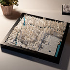 The Chicago 3D Map features a 3D-printed cityscape in a black frame with white buildings and blue water areas, displayed on a white surface with a green plant nearby—an eye-catching wall art piece for any space.