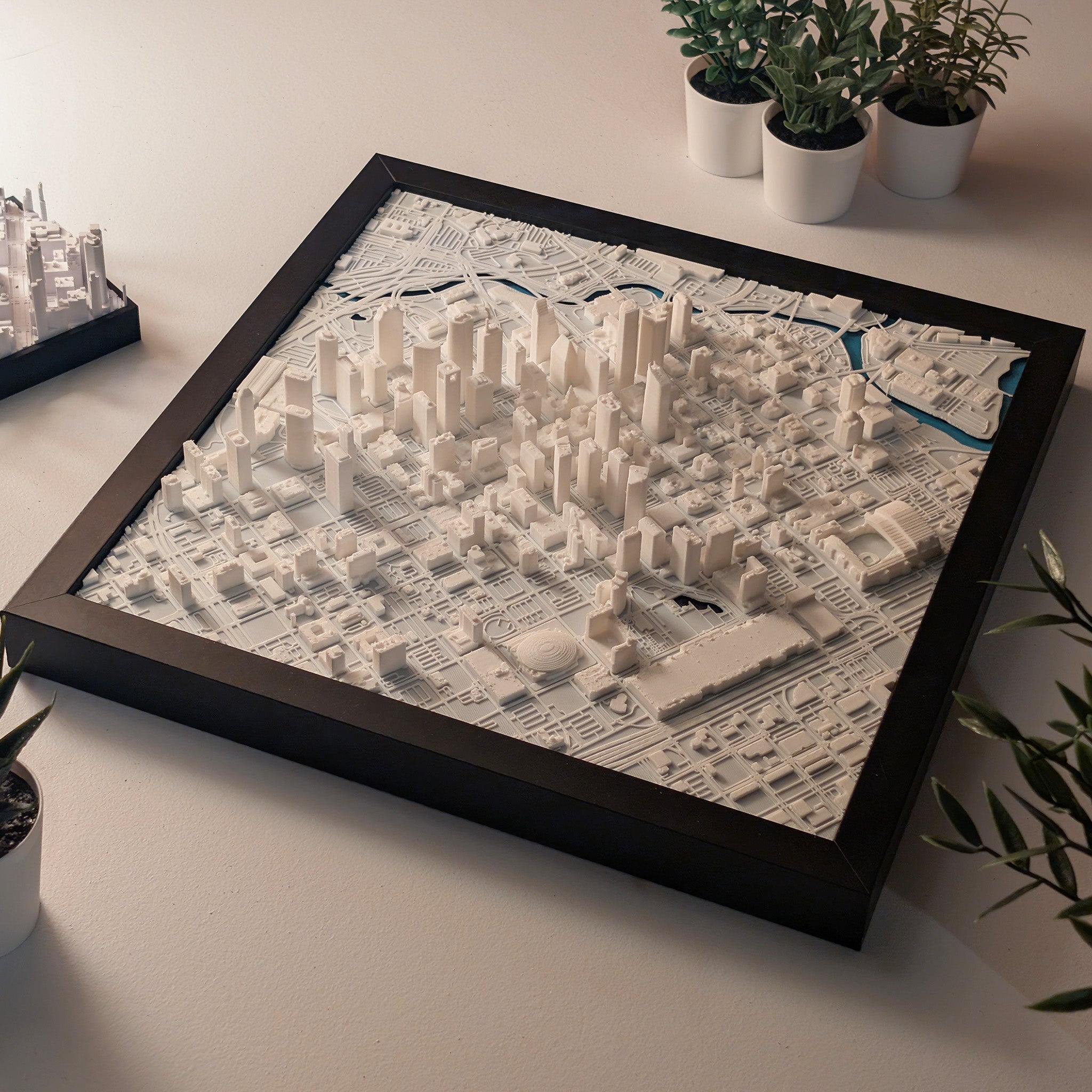 A framed, white Houston 3D Map featuring detailed 3D-printed buildings and streets sits on a table, surrounded by small potted plants.