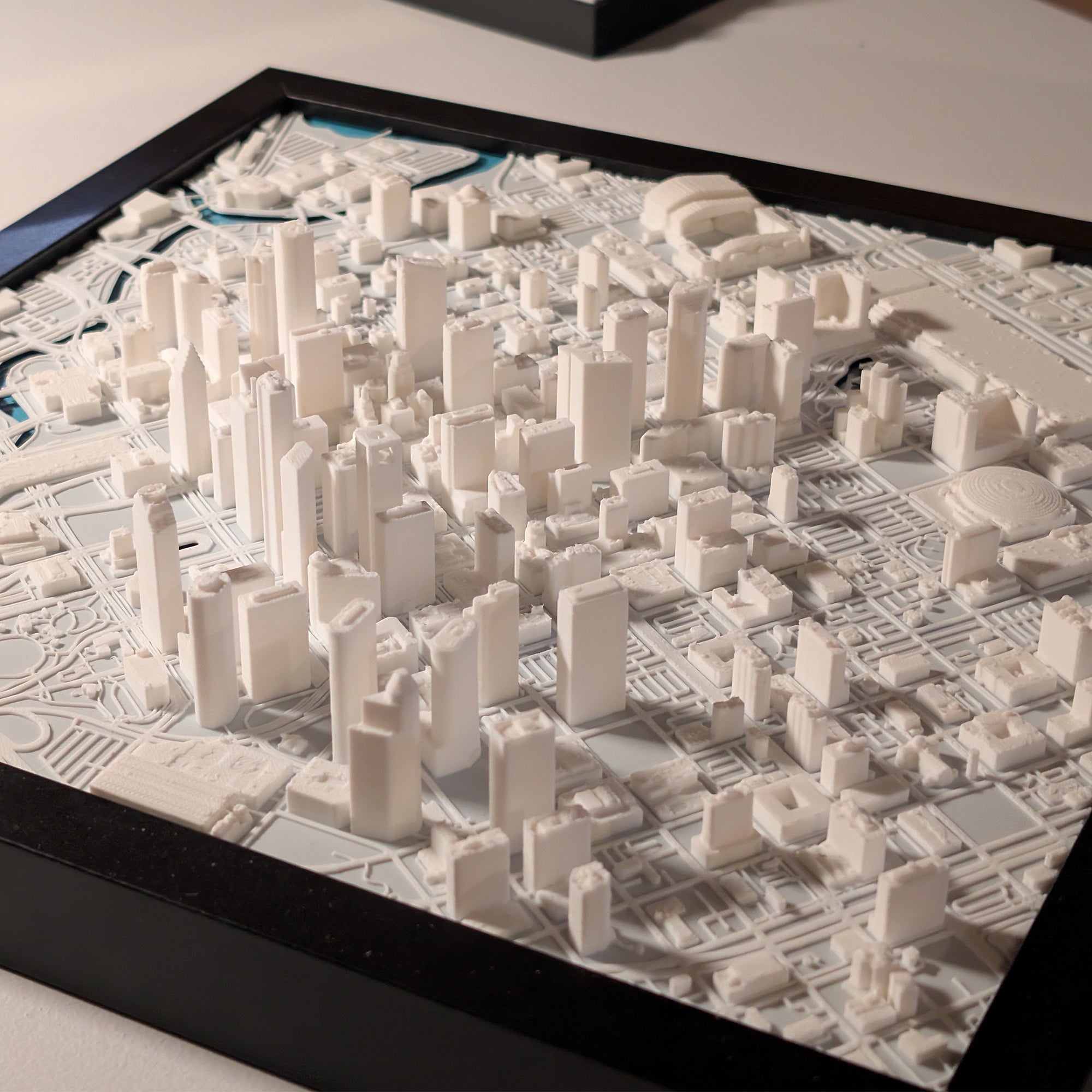 The Houston 3D Map is a detailed white 3D-printed city model featuring high-rise buildings and streets, mounted in a black-framed base for display.