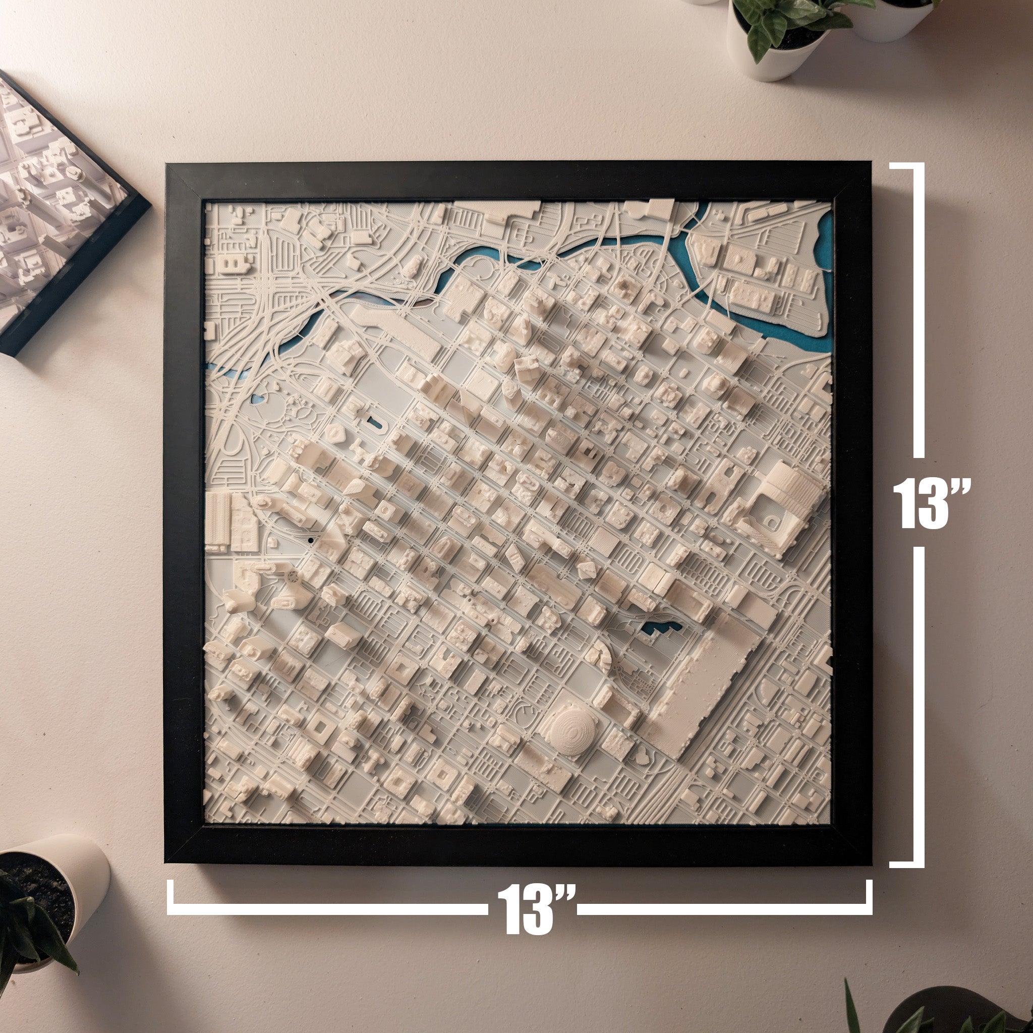 A framed, white Houston 3D Map measuring 13x13 inches hangs on the wall, showcasing detailed buildings, streets, and blue water sections. Small potted plants are placed nearby.