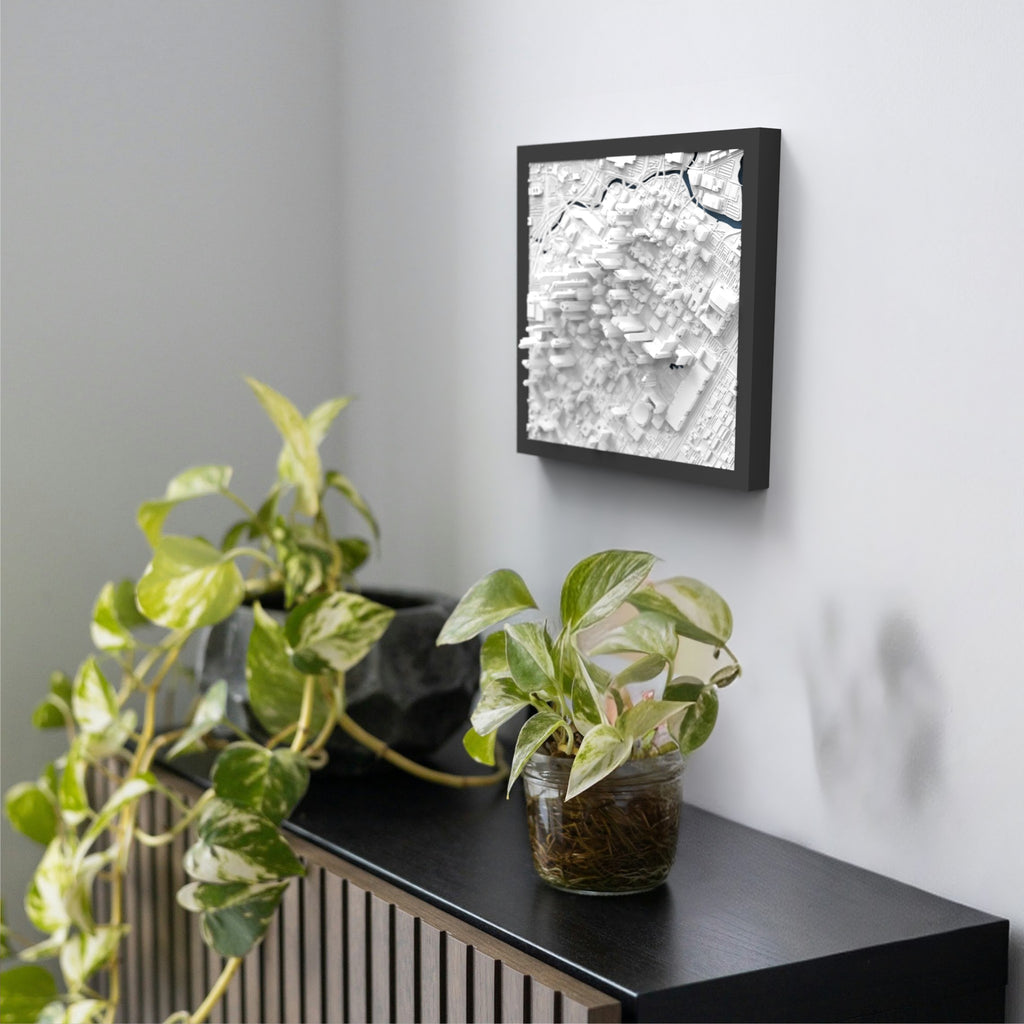 A black shelf with two potted plants sits against a white wall; above it hangs a framed Houston 3D Map artwork featuring abstract geometric shapes in shades of white and gray.