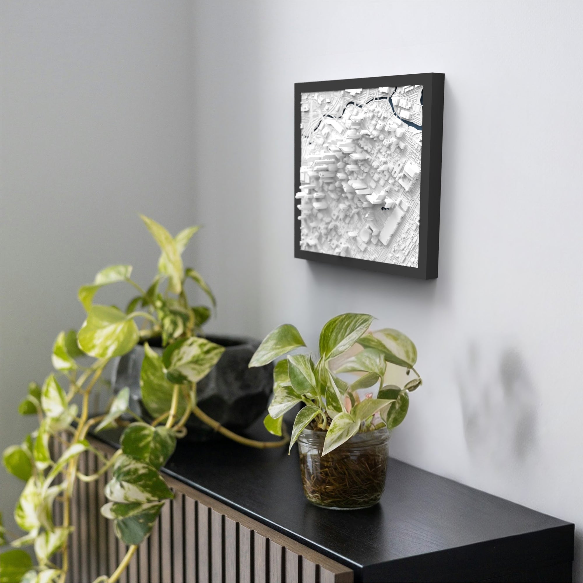 A black shelf with two potted plants sits against a white wall; above it hangs a framed Houston 3D Map artwork featuring abstract geometric shapes in shades of white and gray.