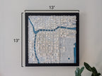 The Chicago 3D Map, a framed wall art measuring 13x13, features white buildings and blue water details for a modern look. Displayed on the wall, it’s complemented by a green plant in the bottom right corner.