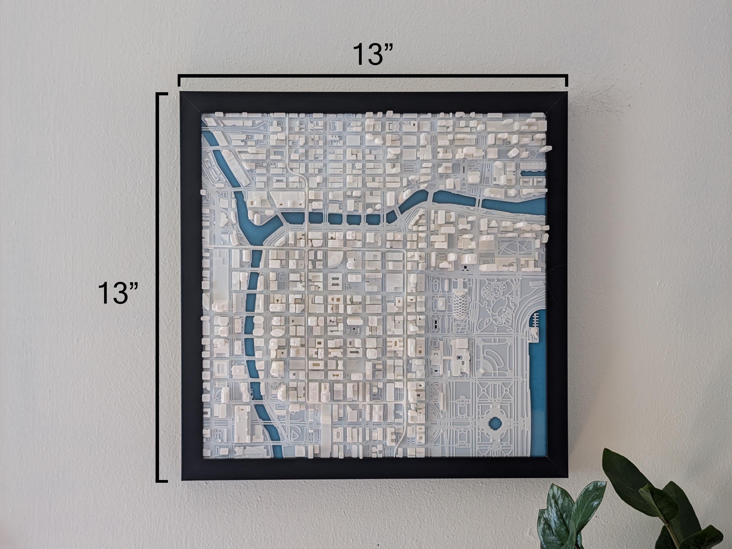 The Chicago 3D Map, a framed wall art measuring 13x13, features white buildings and blue water details for a modern look. Displayed on the wall, it’s complemented by a green plant in the bottom right corner.