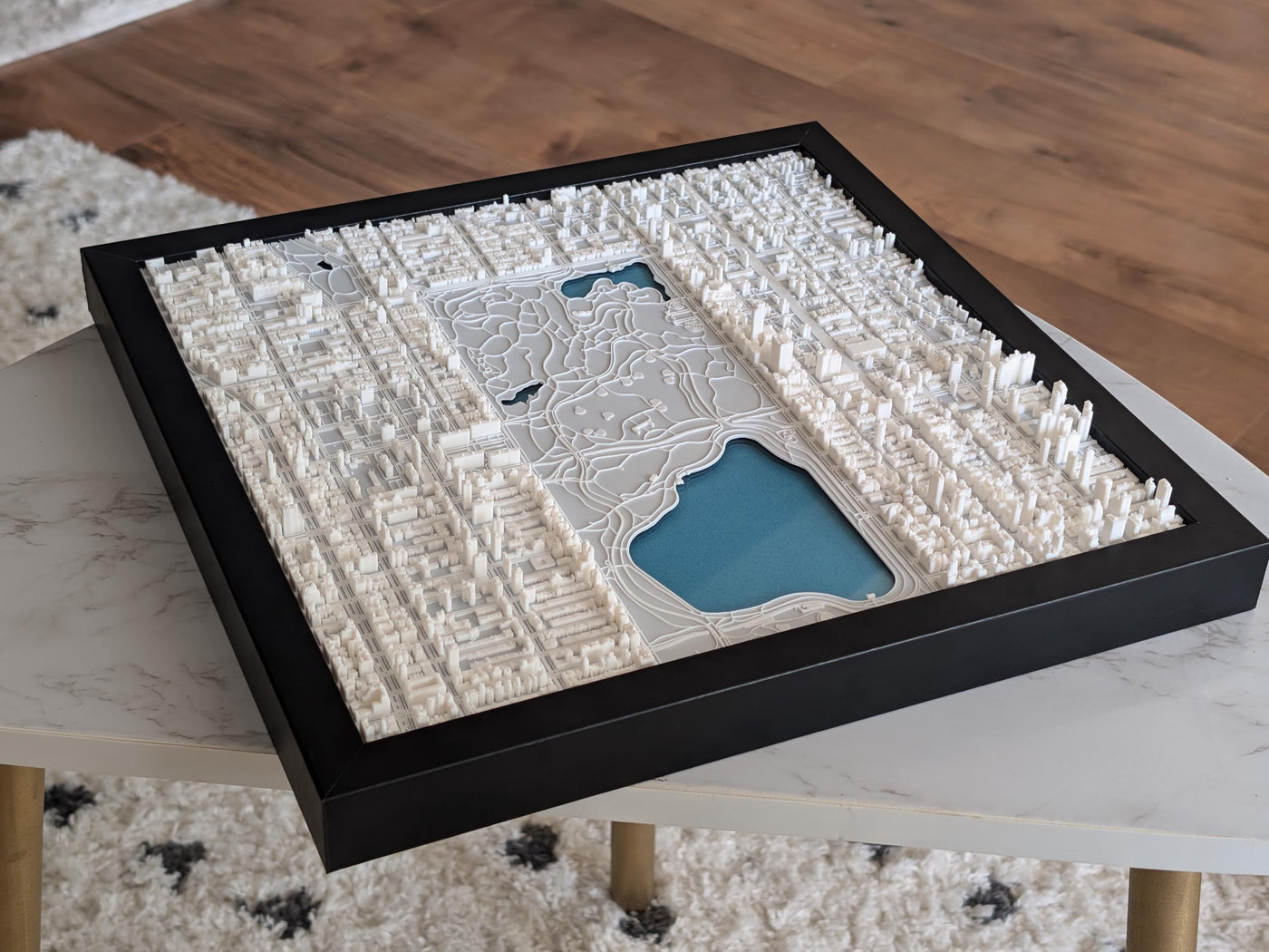 A framed 3D printed map of the northern half of Central Park, featuring white terrains and landmarks against a blue water feature, set in a black frame.